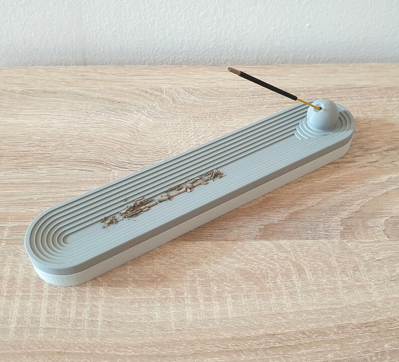 May include: A grey ceramic incense holder with a grooved design and a round knob on the end. The holder is designed to catch ash from incense sticks.