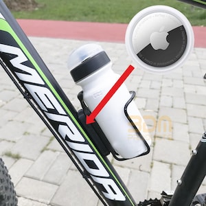 Può includere: Una borraccia bianca con tappo nero è fissata in una gabbia di metallo nero attaccata a un telaio di bicicletta nero e verde. Un AirTag Apple è mostrato nell'angolo in alto a destra.