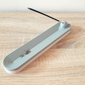 May include: Grey ceramic incense holder with a grooved design and a small round top for holding incense sticks. A black incense stick is resting on the top of the holder.