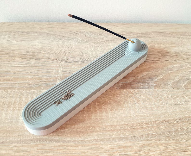 May include: Grey ceramic incense holder with a grooved design and a small round top for holding incense sticks. A black incense stick is resting on the top of the holder.