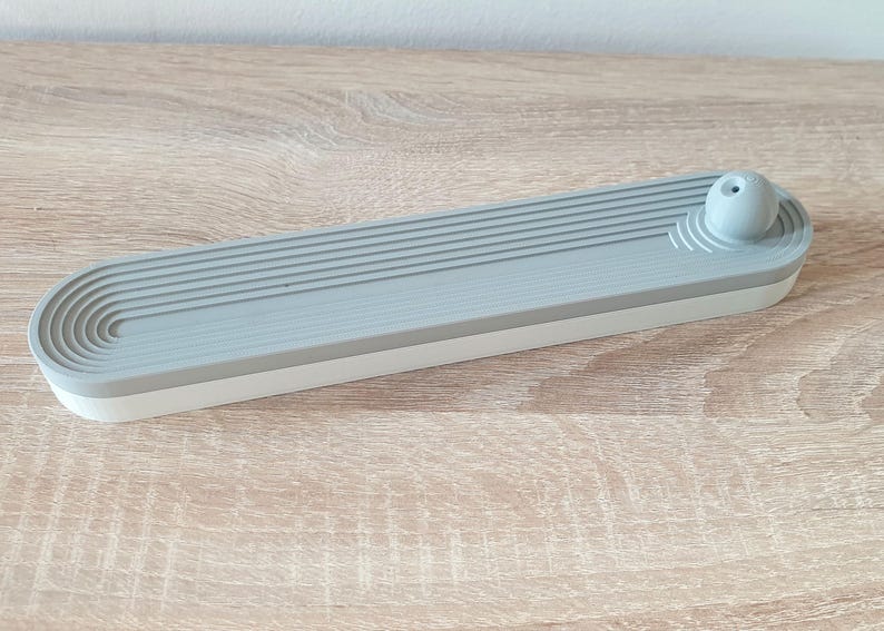 May include: A grey and white ceramic incense holder with a round, grey top. The holder has a series of grooves on the top and a white base.