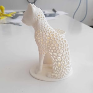 May include: White 3D printed cat figurine with a hollow interior. The cat is sitting with its tail curled around its body. The figurine is made of a lattice-like material.