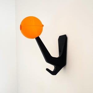 May include: A black wall-mounted hook with an orange sphere on the end. The hook is designed to hold items like coats, hats, or bags.