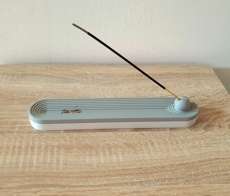 May include: A grey ceramic incense holder with a white base and a round, grey incense stick holder. The incense stick is black and is resting on the holder.
