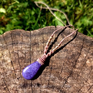 Peut inclure: Un collier artisanal avec un pendentif en pierre précieuse violette en forme de goutte d'eau. Le pendentif est suspendu à un cordon tressé délicat avec des perles dorées. Le collier repose sur une surface en bois vieilli, avec un fond de feuillage vert.