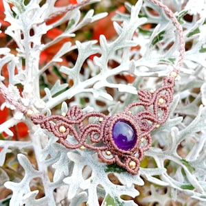 Pode incluir: Um colar de macramé artesanal com uma pedra preciosa central roxa escura. O colar tem um cordão castanho com detalhes intrincados de nós e pequenas contas douradas. O colar é exibido em um fundo de folhagem verde prateada.