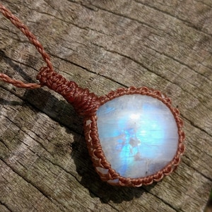 May include: A pendant featuring a smooth, translucent moonstone with a blue iridescent sheen, encased in a brown macrame cord setting. The stone is square-shaped with rounded edges and is attached to a matching brown braided cord necklace.
