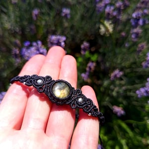 Può includere: Un bracciale in macramè nero con una pietra preziosa rotonda e iridescente dai toni dorati e grigi, arricchito da piccole perline color argento integrate nell'intricato motivo a cordoncino annodato che si estende lungo il cinturino.