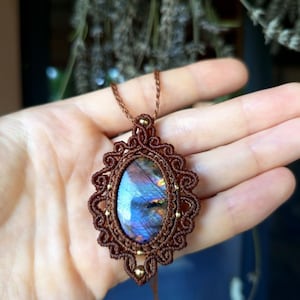 Peut inclure: Un pendentif en pierre de labradorite ovale serti dans un cordon en macramé marron complexe. La pierre présente des reflets irisés bleus et violets. Le sertissage comporte des boucles décoratives et de petites perles métalliques.