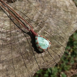 Pode incluir: Um pingente de pedra preciosa verde-claro em forma de lágrima, preso em uma rede de cordão de macramê marrom. O pingente está fixado a um colar de cordão marrom trançado fino que sobe a partir da pedra, com um nó no topo.