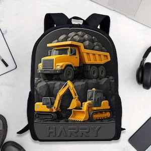 May include: Black backpack featuring a yellow dump truck and construction vehicles on a rocky background. The backpack has the name "HARRY" printed on the bottom. The backpack is surrounded by a laptop, headphones, a phone, a notebook, and shoes.