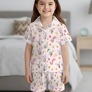 Puede incluir: Una ni&ntilde;a peque&ntilde;a con un conjunto de pijama rosa claro con un estampado repetido de conejitos de Pascua, huevos, zanahorias y flores. La parte superior de manga corta y los pantalones cortos tienen ribetes blancos. La ni&ntilde;a est&aacute; sonriendo.