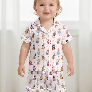 May include: A light pink pajama set for a toddler, featuring a short-sleeved button-up shirt and matching shorts. The set is adorned with a repeating pattern of cartoon bunnies in various colors and designs. White trim outlines the collar, cuffs, and edges.