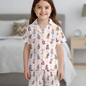 May include: White pajama set with a pattern of cartoon bunnies and princesses. The short-sleeved top and shorts feature a button-down shirt style with a drawstring waist. The pajamas are designed for children.