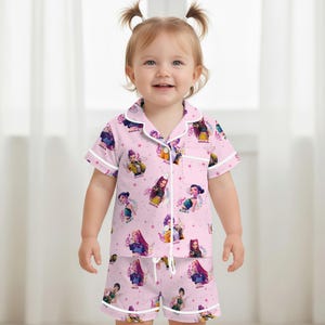 Peut inclure: Ensemble de pyjama rose pour enfant avec un motif r&eacute;p&eacute;t&eacute; de personnages de dessins anim&eacute;s, de notes de musique et d'&eacute;toiles. Le haut &agrave; manches courtes et le short ont une bordure blanche. Les noms "MIRA" et "ZOEY" sont visibles.