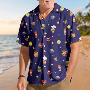 May include: A dark blue short-sleeved button-down shirt featuring an all-over print of various Muppet characters including Kermit the Frog, Fozzie Bear, Animal, and the Swedish Chef, interspersed with small yellow stars across the fabric.