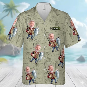 May include: A short-sleeved button-down shirt with a light green background featuring a repeating pattern of fish hooks and cartoon illustrations of a fisherman holding a fish. The name "Jack" is printed on the shirt.
