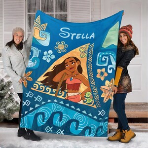 May include: A blue fleece blanket featuring a central triangular graphic of the character Moana, the name Stella in white script, and various tropical floral and wave patterns in shades of blue, orange, and yellow across the fabric surface.