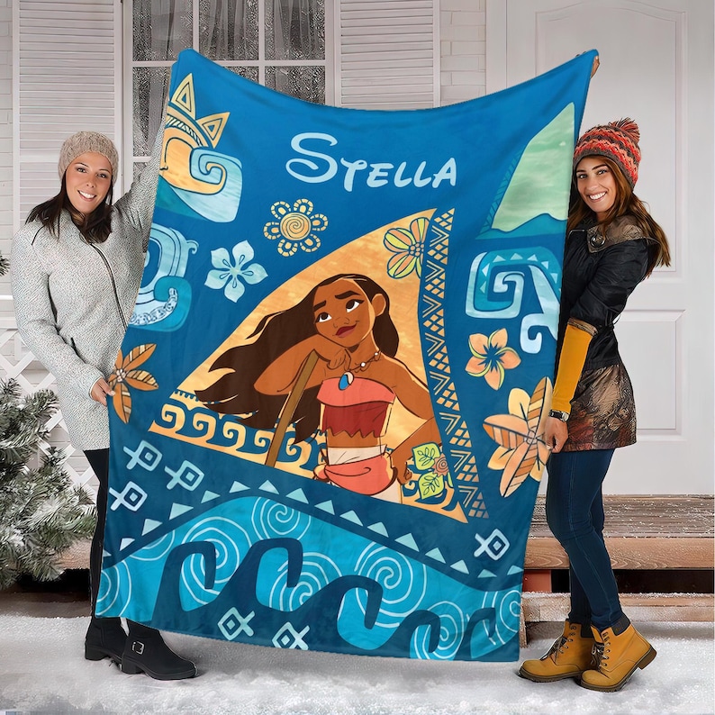 May include: A blue fleece blanket featuring a central triangular graphic of the character Moana, the name Stella in white script, and various tropical floral and wave patterns in shades of blue, orange, and yellow across the fabric surface.