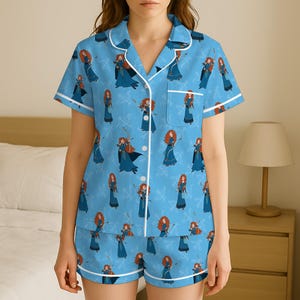 Op de afbeelding: Lichtblauwe pyjamaset met een herhalend patroon van een roodharig personage in een blauwe jurk. De top met korte mouwen en de shorts hebben witte biezen. De pyjama is gemaakt van een zacht materiaal.
