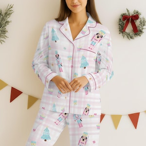 May include: A two-piece pajama set featuring a white and pink plaid pattern. The pajamas are adorned with a repeating print of nutcrackers, Christmas trees, and cupcakes. The set has pink trim and a button-down shirt with a notched collar.