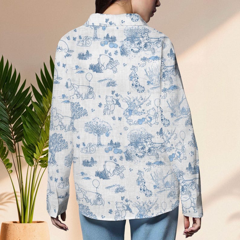 May include: White button-down shirt with a blue toile pattern featuring Winnie the Pooh and friends. The long-sleeved shirt has a classic collar and is made of a lightweight fabric. The design includes trees, balloons, and characters.