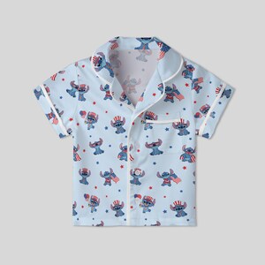 May include: Light blue pajama shirt featuring a repeating pattern of a cartoon character in patriotic attire. The short-sleeved shirt has white trim and a chest pocket. The cartoon character is depicted with an American flag and a patriotic hat.