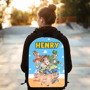 Pode incluir: Mochila azul com o nome "HENRY" em letras amarelas. O design apresenta personagens de desenhos animados, incluindo um cowboy e um dinossauro, num fundo de c&eacute;u azul. Ideal para a escola.