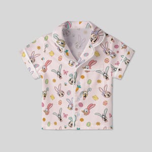 Puede incluir: Una camisa de pijama rosa claro de manga corta con un estampado tem&aacute;tico de Pascua. El estampado incluye conejos de dibujos animados, huevos, mariposas y flores. La camisa tiene cuello y cierre de botones.