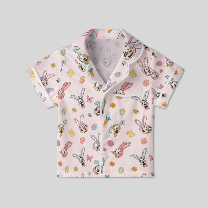 Puede incluir: Una camisa de pijama rosa claro de manga corta con un estampado tem&aacute;tico de Pascua. El estampado incluye conejos de dibujos animados, huevos, mariposas y flores. La camisa tiene cuello y cierre de botones.