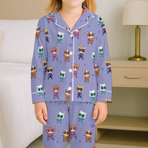 Puede incluir: Un conjunto de pijama de color lila claro con un patr&oacute;n repetido de personajes de gatos de dibujos animados en varios colores y poses. La parte superior de manga larga y los pantalones tienen ribetes blancos y formas de estrella.