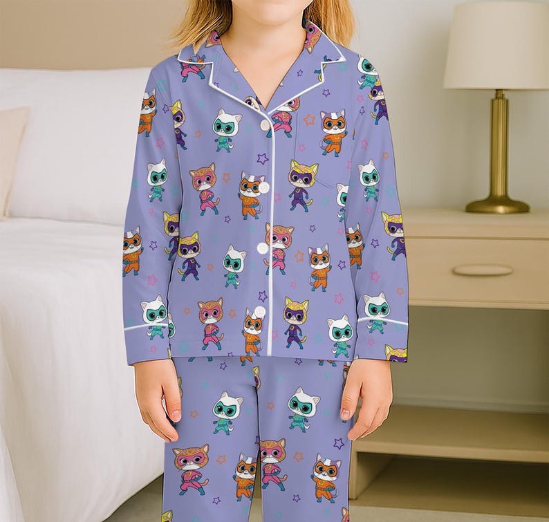 Puede incluir: Un conjunto de pijama de color lila claro con un patr&oacute;n repetido de personajes de gatos de dibujos animados en varios colores y poses. La parte superior de manga larga y los pantalones tienen ribetes blancos y formas de estrella.