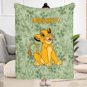 May include: A personalized blanket featuring a cartoon lion cub in shades of yellow and orange, with the name "HENRY" in gold letters. The background is a light green with a leaf pattern.