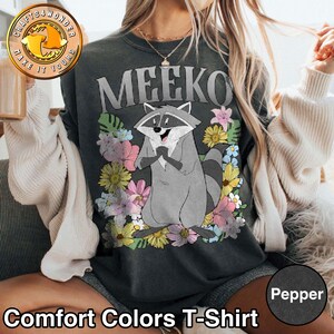 Pu&ograve; includere: Una maglietta Comfort Colors grigio scuro con la scritta "MEEKO" sopra un personaggio di procione dei cartoni animati circondato da fiori colorati. Il procione &egrave; grigio con una coda a strisce bianche e nere. La maglietta ha anche il testo "Comfort Colors T-Shirt" in basso.