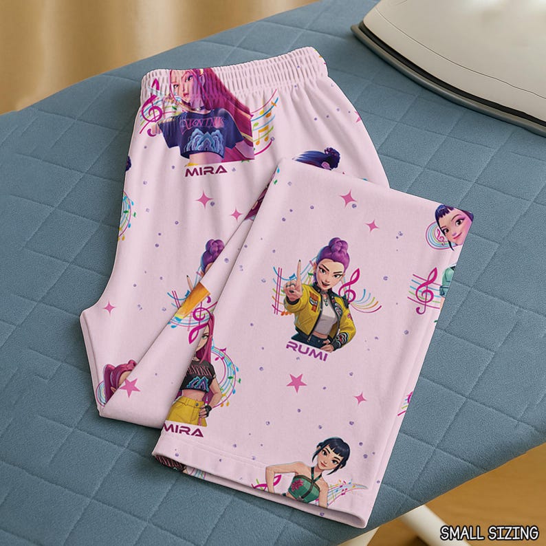 May include: Pink pajama set featuring illustrations of animated female characters, musical notes, and stars. The pajama pants and top are decorated with the names "MIRA" and "RUMI". The set is made of a soft, lightweight fabric.