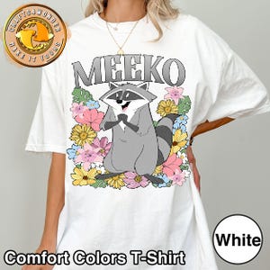 Pu&ograve; includere: T-shirt bianca Comfort Colors con un procione dei cartoni animati di nome Meeko, circondato da fiori colorati. Il procione &egrave; grigio con una maschera nera e una coda a strisce. La parola "MEEKO" &egrave; stampata sopra il procione in lettere grigie.