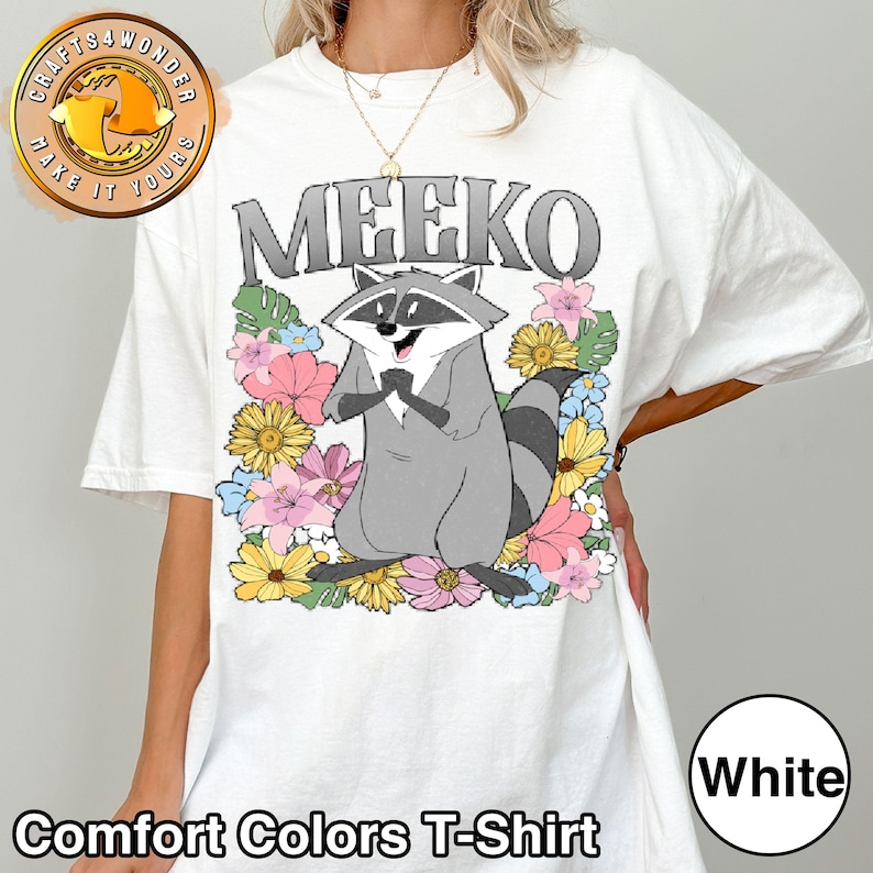 Pu&ograve; includere: T-shirt bianca Comfort Colors con un procione dei cartoni animati di nome Meeko, circondato da fiori colorati. Il procione &egrave; grigio con una maschera nera e una coda a strisce. La parola "MEEKO" &egrave; stampata sopra il procione in lettere grigie.