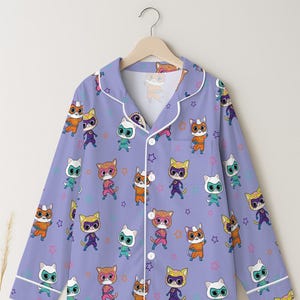 Puede incluir: Una camisa de pijama lila claro con un patr&oacute;n repetido de superh&eacute;roes de dibujos animados de gatos en varios colores. El pijama tiene ribetes y botones blancos, y est&aacute; colgado de una percha de madera.