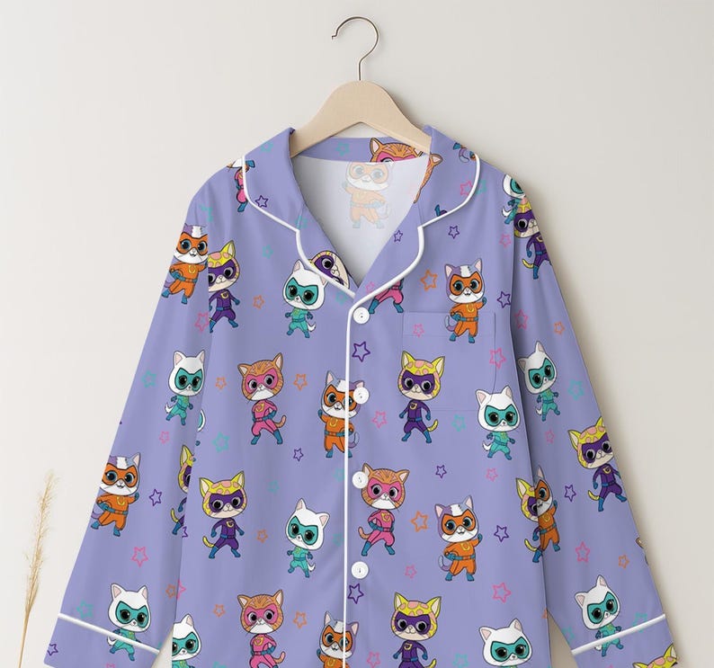 Puede incluir: Una camisa de pijama lila claro con un patr&oacute;n repetido de superh&eacute;roes de dibujos animados de gatos en varios colores. El pijama tiene ribetes y botones blancos, y est&aacute; colgado de una percha de madera.