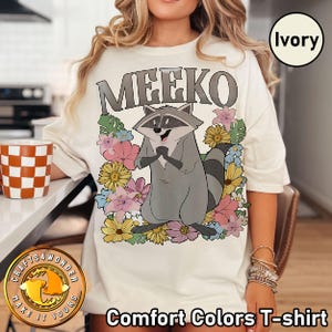 Pu&ograve; includere: T-shirt Comfort Colors color avorio con un procione dei cartoni animati di nome Meeko, circondato da fiori colorati. Il procione &egrave; grigio con una coda a strisce ed &egrave; posizionato al centro della maglietta.