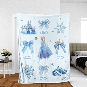 Op de afbeelding: Lichtblauwe deken met aquarel illustraties van een kasteel, sneeuwvlokken, strikken, een kroon en een berg. De deken heeft een afbeelding van een prinses in een blauwe jurk. De naam "Stella" is in lichtblauw geschreven.