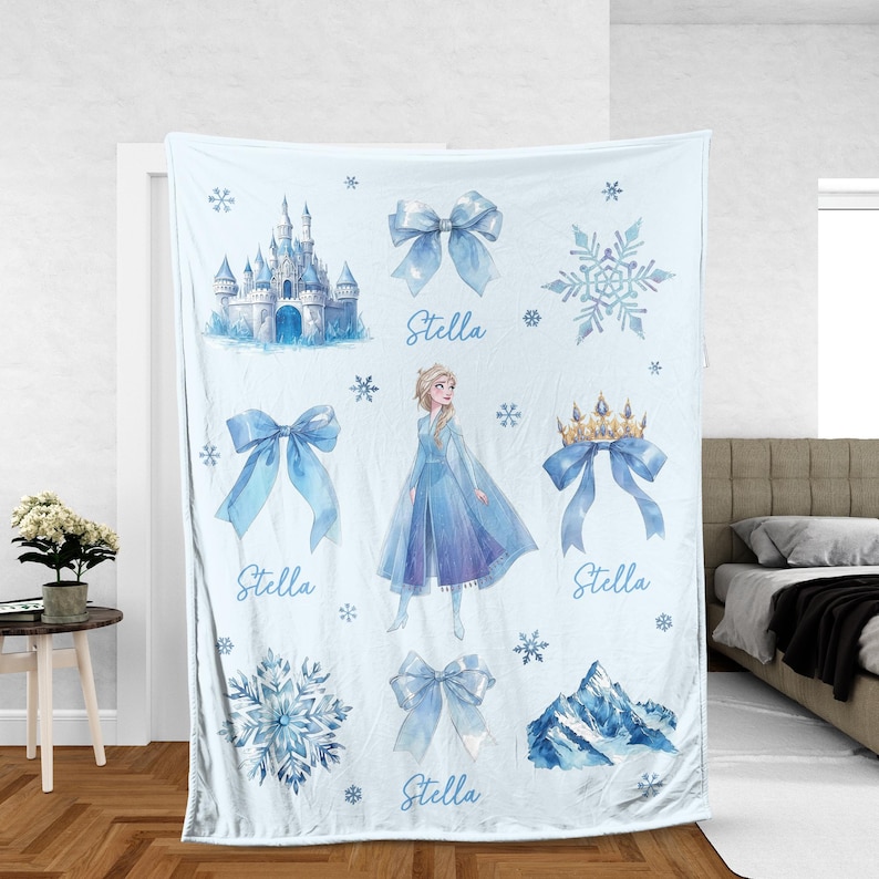 Op de afbeelding: Lichtblauwe deken met aquarel illustraties van een kasteel, sneeuwvlokken, strikken, een kroon en een berg. De deken heeft een afbeelding van een prinses in een blauwe jurk. De naam "Stella" is in lichtblauw geschreven.