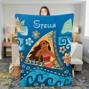 May include: A blue fleece blanket featuring a central triangular graphic of a cartoon character with long brown hair, wearing a red top and skirt. The blanket displays the name Stella in white text, surrounded by floral and wave patterns.