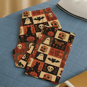 Peut inclure: Un legging sur le thème d'Halloween avec un motif répété de citrouilles, de fantômes, de chauves-souris, de crânes et de livres. Le legging est dans un design patchwork avec des carrés noirs, orange et crème. Il est plié sur une planche à repasser bleue.