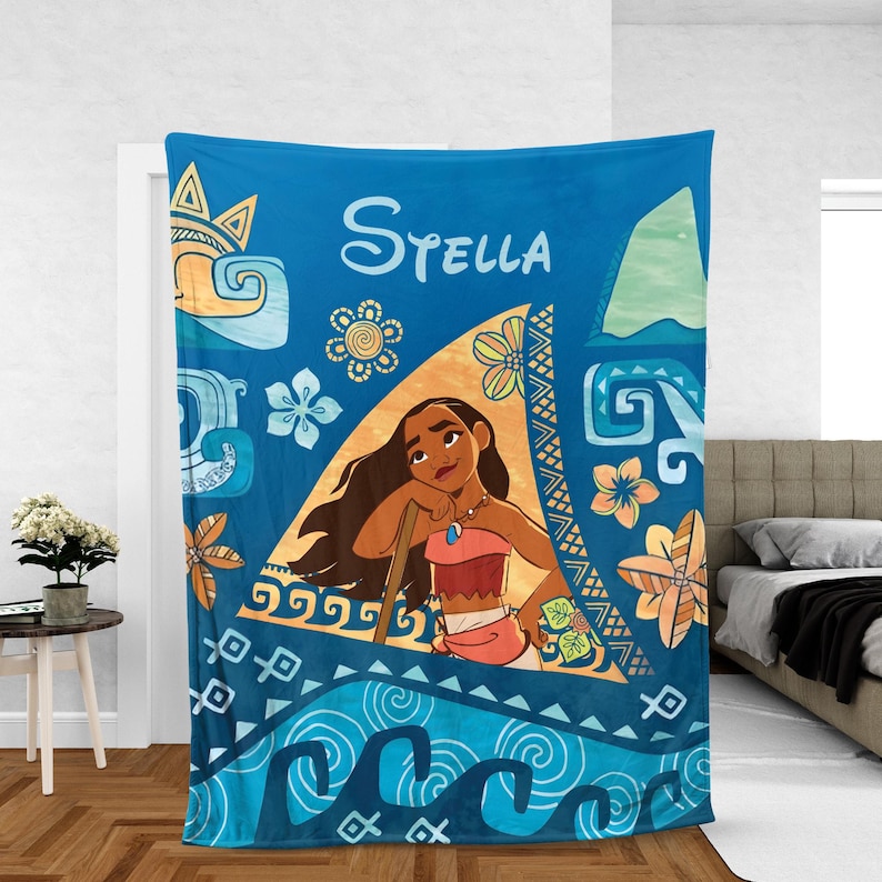 May include: A blue fleece blanket featuring a graphic of the character Moana centered within a triangular frame. The design includes the name Stella in white script, stylized ocean waves, tropical flowers, and geometric patterns in shades of blue, orange, and yellow.