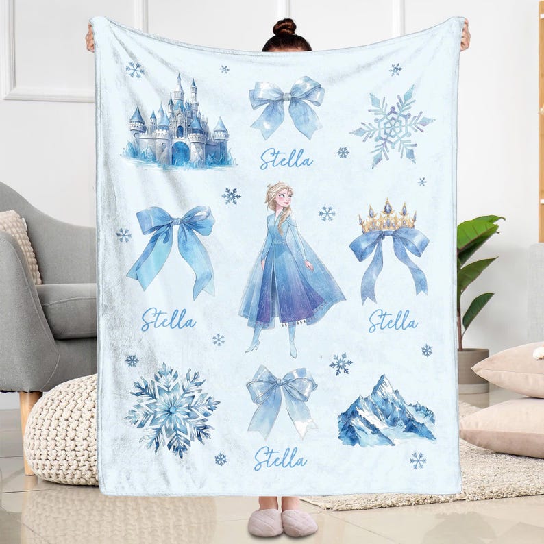 Op de afbeelding: Een lichtblauwe deken met aquarel illustraties van een kasteel, sneeuwvlokken, strikken, een kroon en een berg. De deken heeft ook een prinses in een blauwe jurk en de naam "Stella".