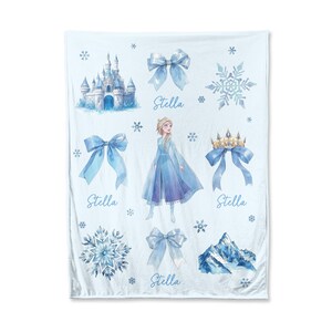 Op de afbeelding: Een lichtblauwe deken met illustraties van een kasteel, sneeuwvlokken, strikken, een berg en een prinses. De naam "Stella" staat in script onder enkele van de afbeeldingen. De deken is wit met een lichtblauw ontwerp.