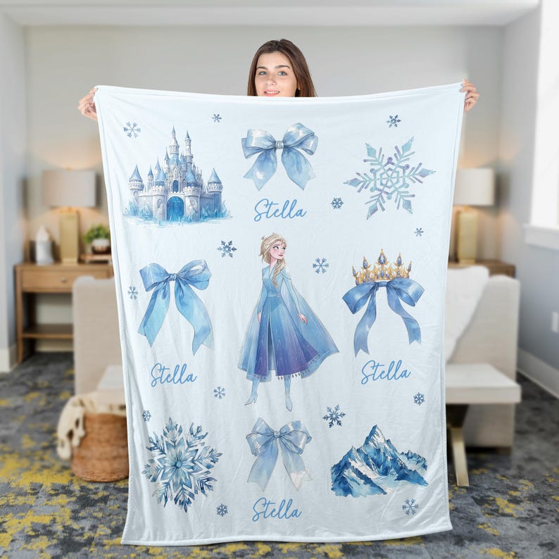 Op de afbeelding: Een lichtblauwe deken met illustraties van een kasteel, sneeuwvlokken, strikken, een kroon en een berg. De deken toont ook een prinses in een blauwe jurk en de naam "Stella" in een sierlijk lettertype.