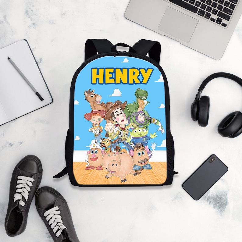 Pode incluir: Uma mochila preta com fundo azul com a palavra "HENRY" em amarelo. A mochila tem um gr&aacute;fico colorido de personagens do filme Toy Story. A mochila &eacute; cercada por t&ecirc;nis pretos, um caderno, um laptop, fones de ouvido e um smartphone.