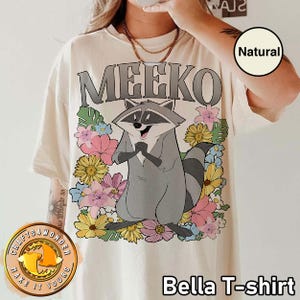 Pu&ograve; includere: T-shirt Bella color crema con un personaggio di procione dei cartoni animati di nome Meeko, circondato da fiori colorati. La parola "MEEKO" &egrave; stampata sopra il procione in lettere grigie. La maglietta ha una vestibilit&agrave; ampia.